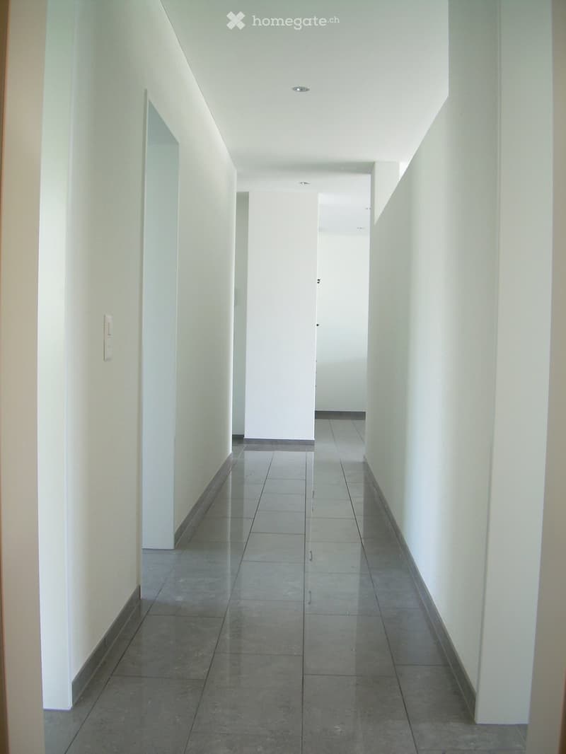 Empty hallway with tiled floor and white walls, featuring a closed door at the end and a light switch on the left wall.