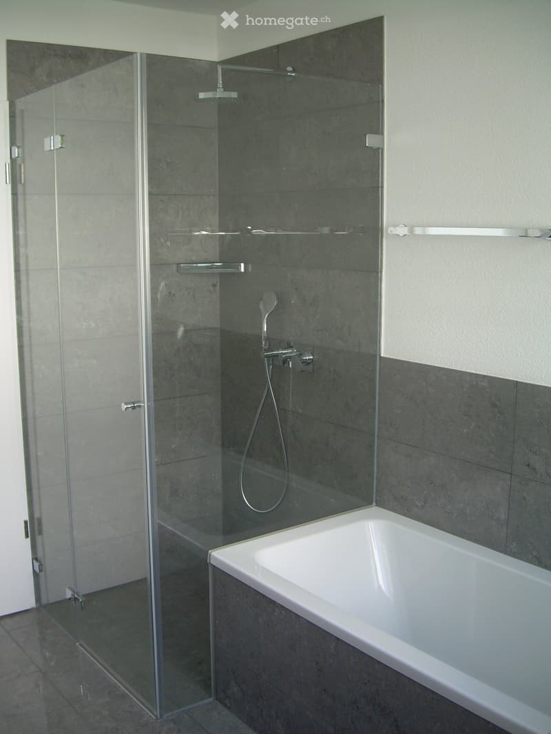 Modern bathroom with a white bathtub, glass-enclosed shower, and grey tiled walls.