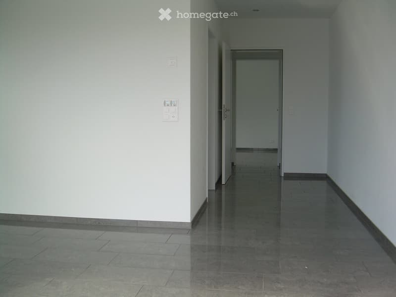 Empty hallway with tiled floor, white walls, and light switches on the left wall. A door is visible at the end of the hallway.