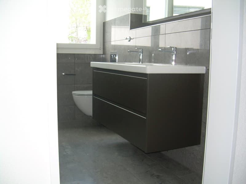 Modern bathroom with a double sink, sleek dark cabinet, chrome faucets, and a large mirror above the sink. The floor and walls are tiled in a dark gray color.