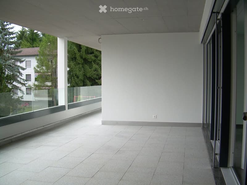Modern balcony with large glass windows, white tiled floor, and a view of greenery and neighboring buildings.