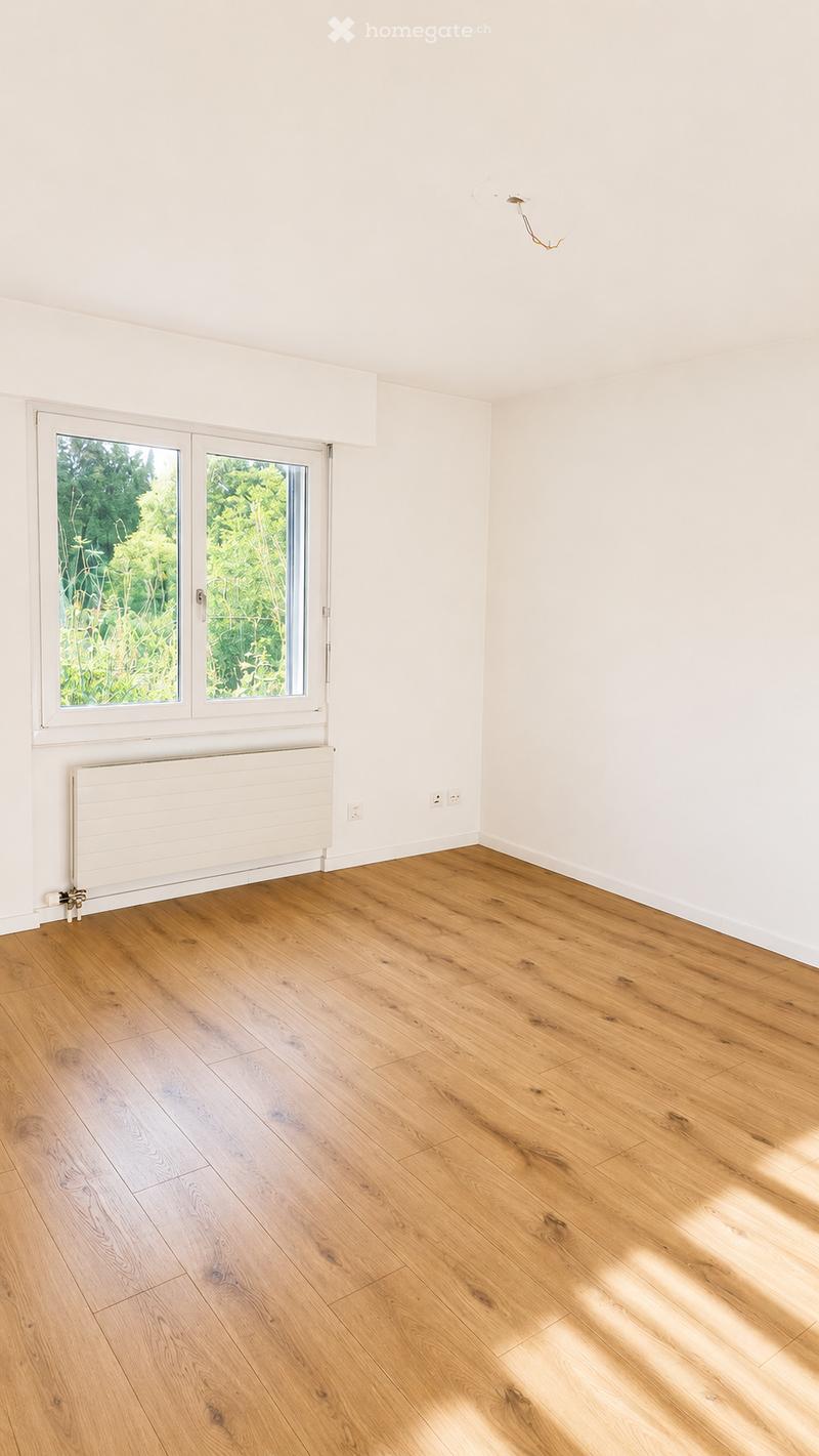 Empty room with light wooden flooring, white walls, a large window with a view of greenery, and a radiator below the window.
