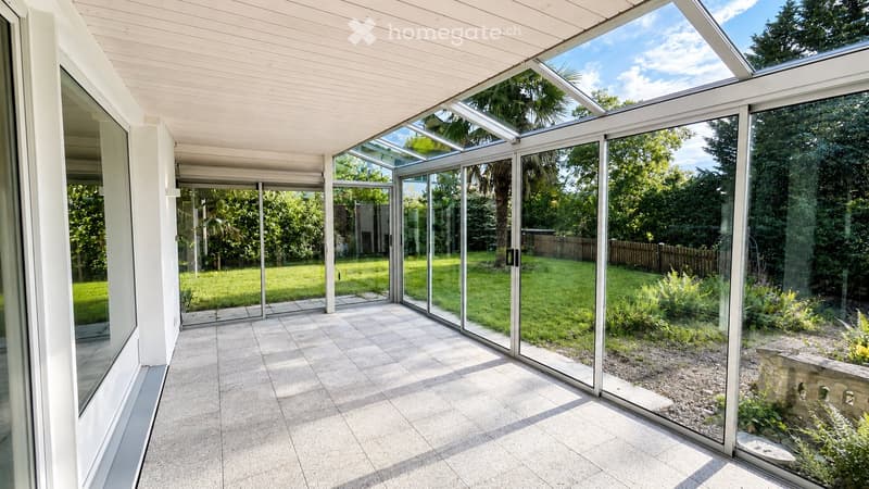 Bright, spacious sunroom with large glass walls and a skylight, overlooking a well-maintained garden with trees and shrubs.