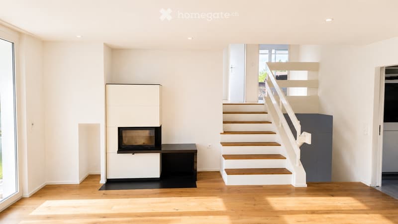 Bright, minimalist hallway with wooden flooring, white walls, and a staircase leading upwards. Features a built-in fireplace with a black surround and a small bench, along with a large window on the left side.