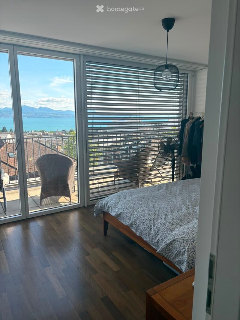 Bright bedroom with wooden flooring, large windows offering a scenic view, a bed with a patterned duvet, a wicker chair, a standing fan, and a wardrobe with hanging clothes.