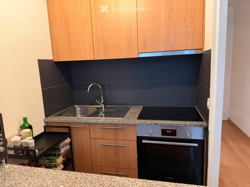 Modern kitchen with light wooden cabinets, granite countertop, stainless steel sink, and Bosch oven. Includes a small cart with cups and a bottle on the counter.