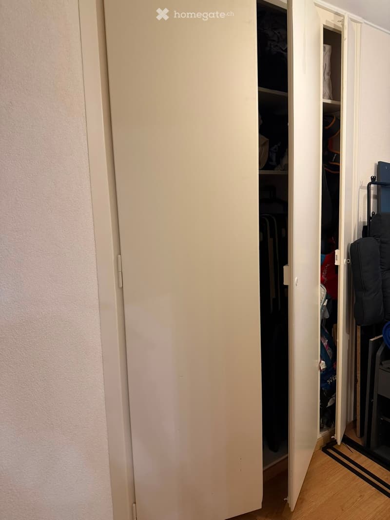 A narrow hallway with a partially open closet door revealing stored items, light-colored walls, and wooden flooring.