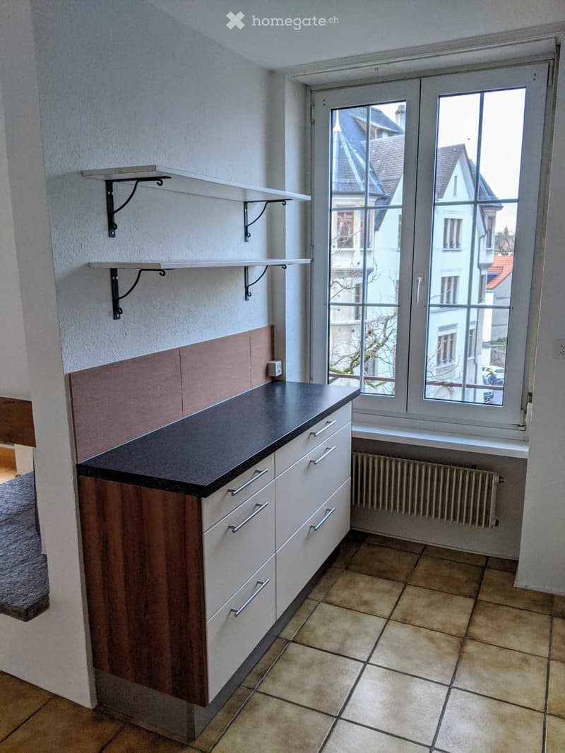 Kitchen. Modern kitchen with white cabinets, dark countertop, built-in drawers, and a radiator beneath the window. The room features tiled flooring and a window with a view of neighboring buildings.