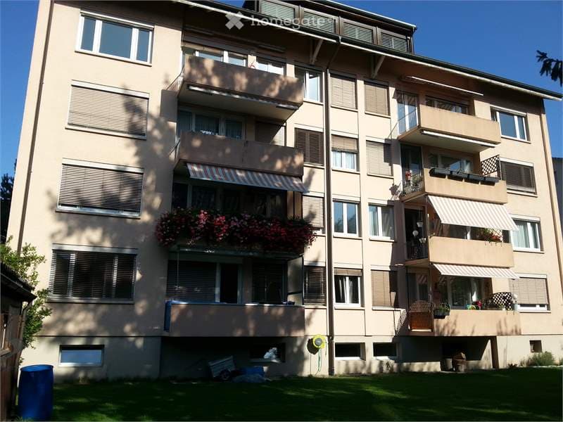 Multi-story residential building with balconies, large windows, and flower boxes. Beige facade with shutters and awnings.