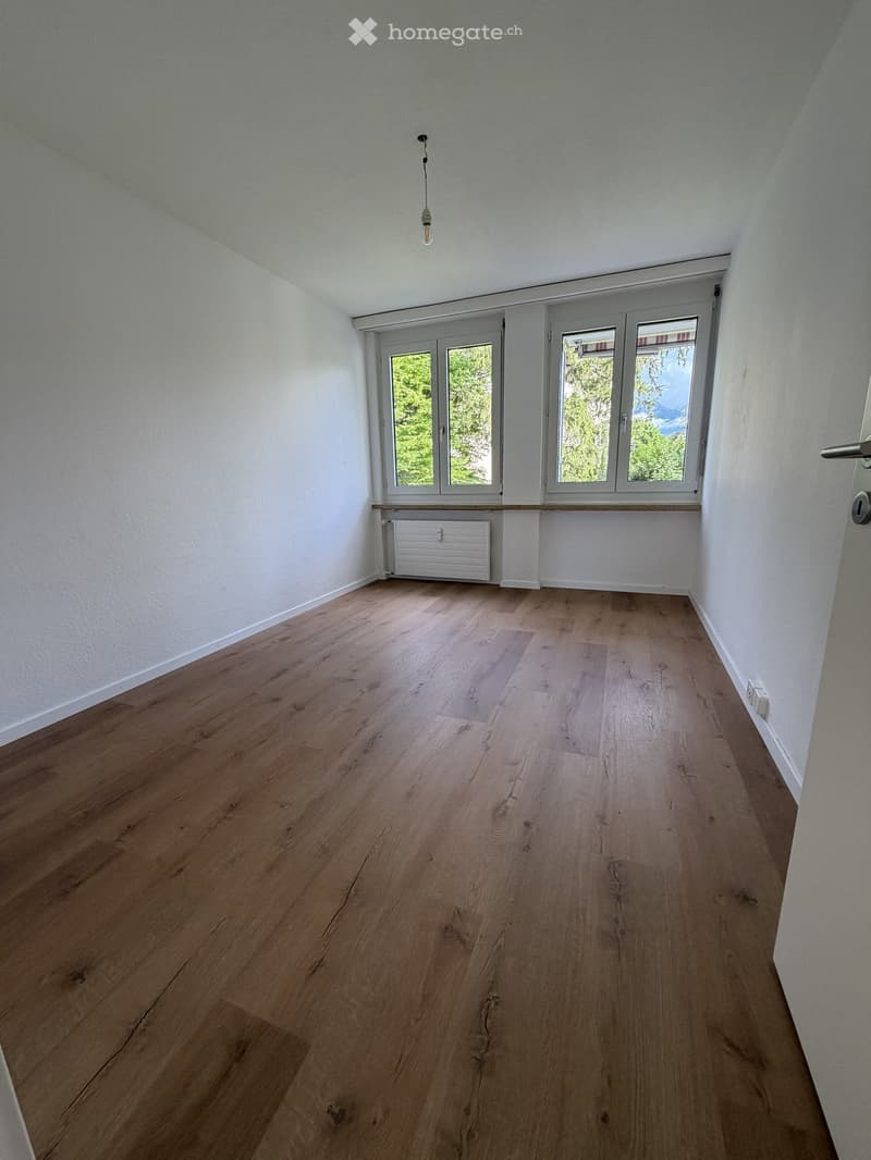 Empty room with light wooden flooring, white walls, and large windows with a view of greenery. A radiator is visible under the windows.