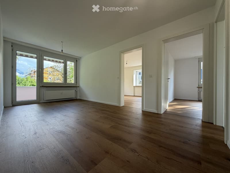 Empty room with light wooden flooring, white walls, large windows, and open doorways leading to other spaces.