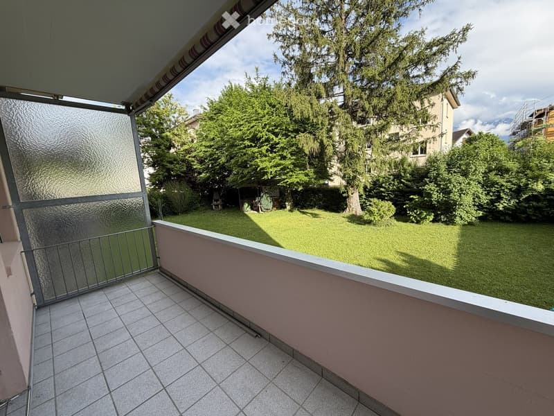Balcony with tiled floor, glass railing, and view of a green lawn and trees.