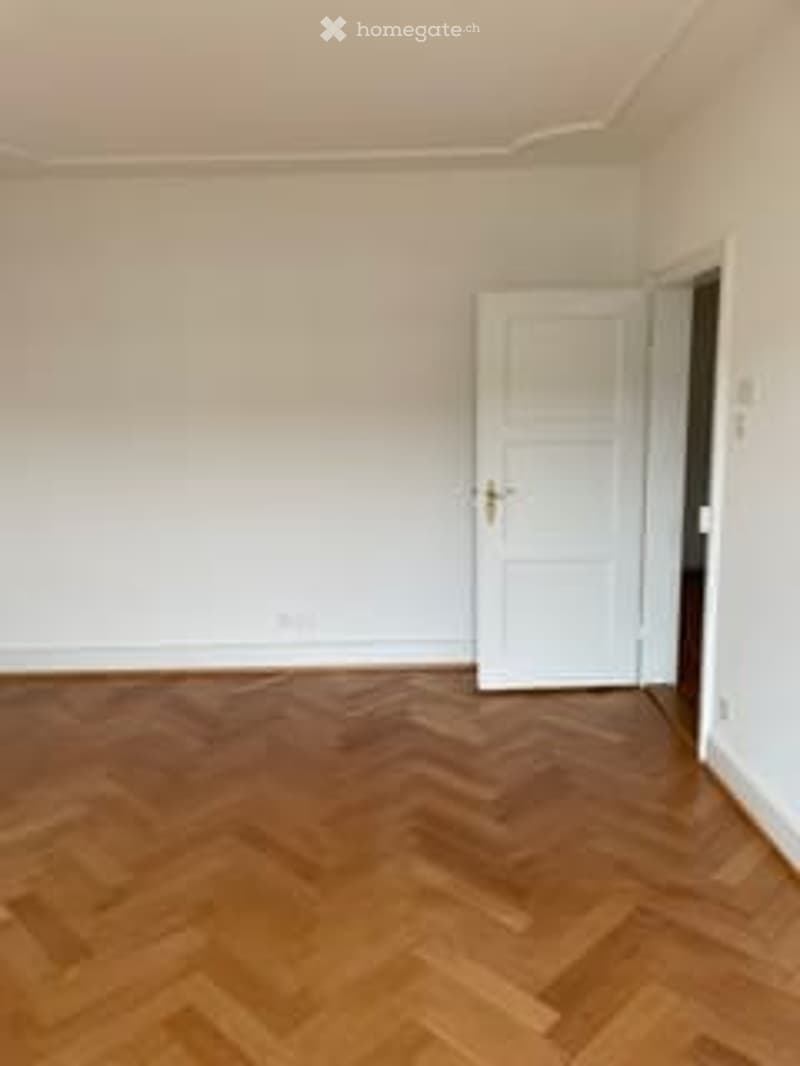 Completely empty room with light wooden herringbone flooring and white walls. A white door is visible on the right side.