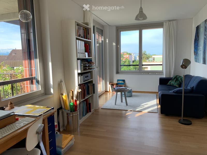 Bright living room with wooden flooring, large windows offering a scenic view, a blue sofa, a small round table, a bookshelf filled with books, a desk with a keyboard, and a floor lamp.