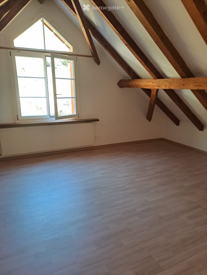 Empty attic room with wooden beams, wooden floor, and large windows allowing natural light.