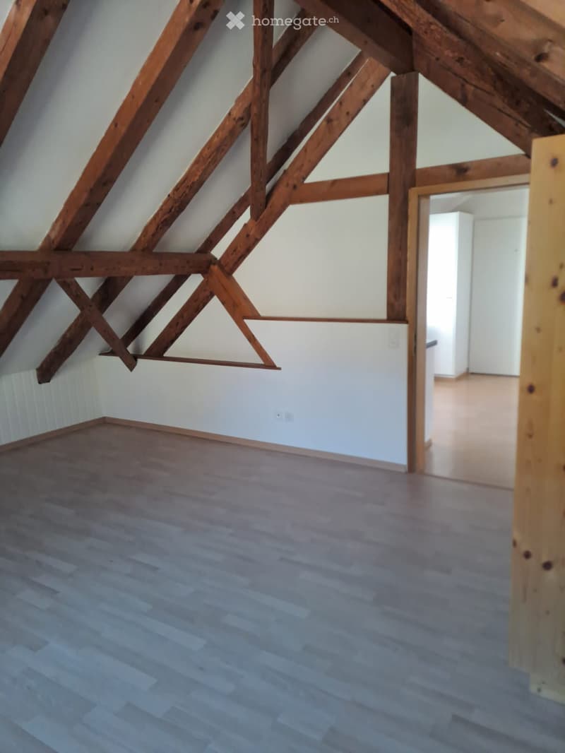 Empty attic space with wooden beams, light-colored walls, and a wooden floor. No furniture or equipment is present.