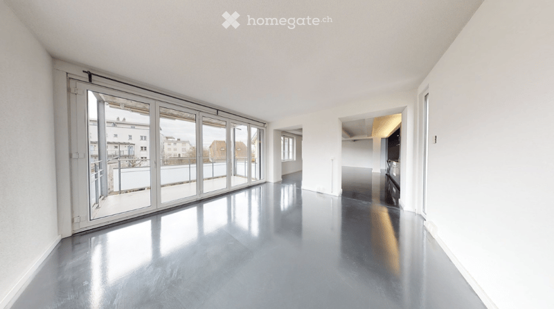 Empty room with large sliding glass doors leading to a balcony, polished concrete floor, white walls, and visible kitchen area in the background.