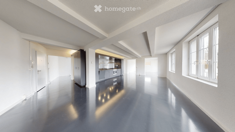 Open-plan unfurnished space with polished concrete floors, white walls, and large windows. The area includes a small kitchen area with cabinets and appliances, but no furniture is present.