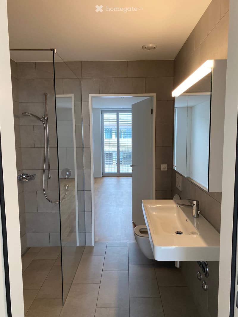 Bathroom. Modern bathroom with a glass-enclosed shower, a white sink with a modern faucet, a toilet, and a large mirror with built-in lighting. The space features tiled walls and floors and an open doorway leading to another room.