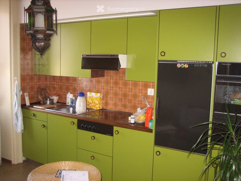 Green kitchen with stainless steel appliances, including an oven, refrigerator, and sink. Countertop has various items including a cutting board, milk, and a box of bread. A wicker table with papers is in the foreground.