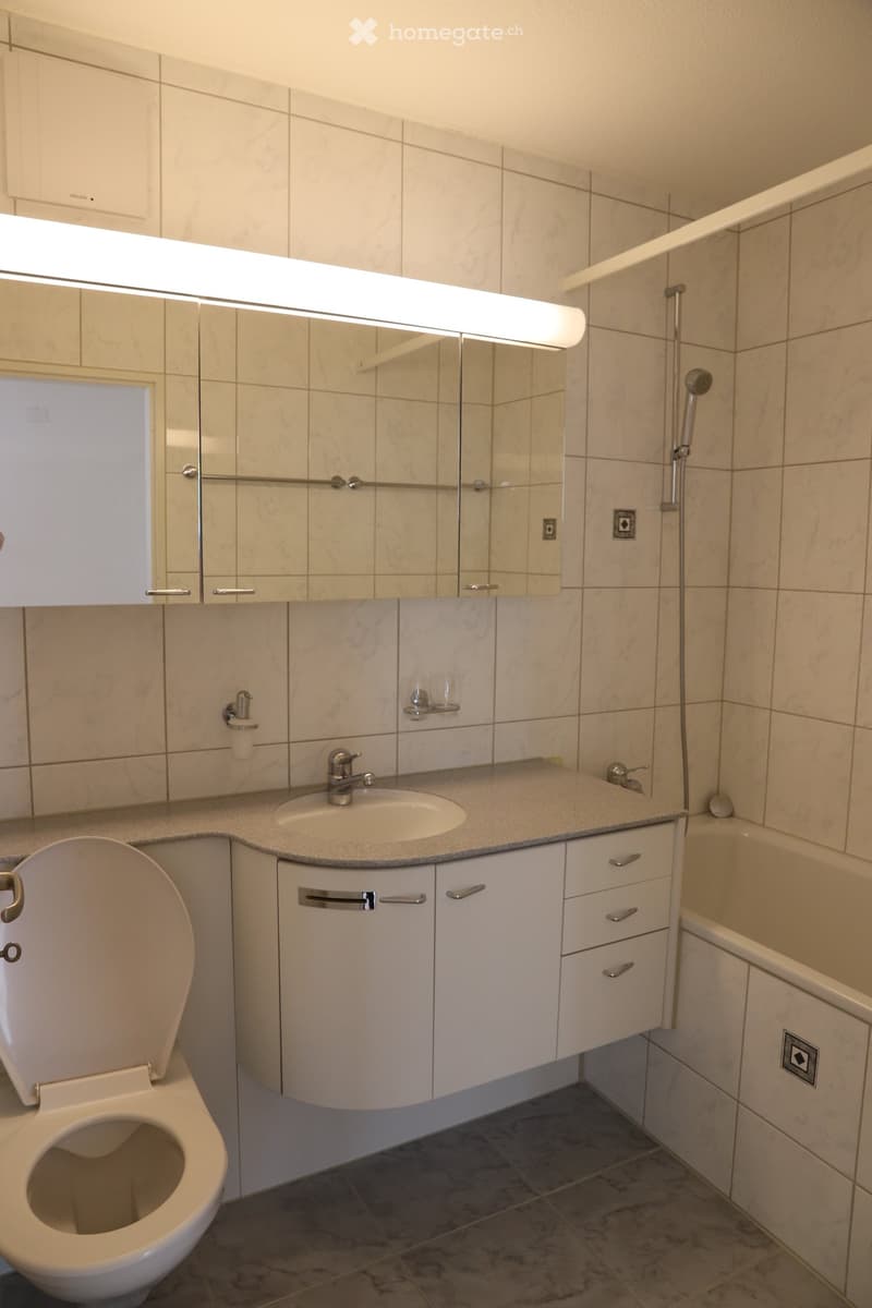 Modern bathroom with a white toilet, sink, and bathtub. Features a large mirror with built-in lighting, a modern faucet, and white cabinets with silver handles.