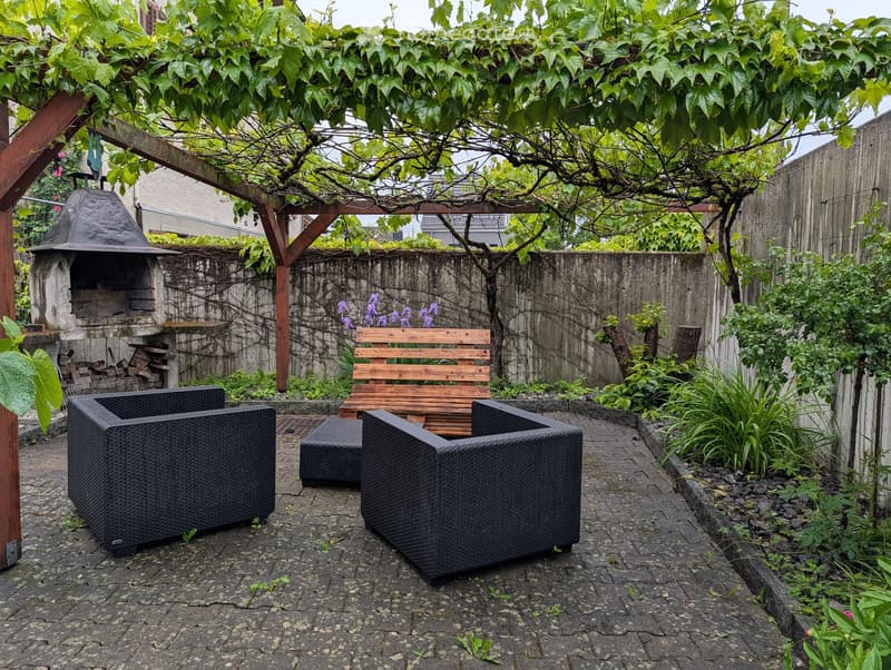 A small outdoor patio with a stone barbecue, wooden bench, wicker chairs, and lush greenery.