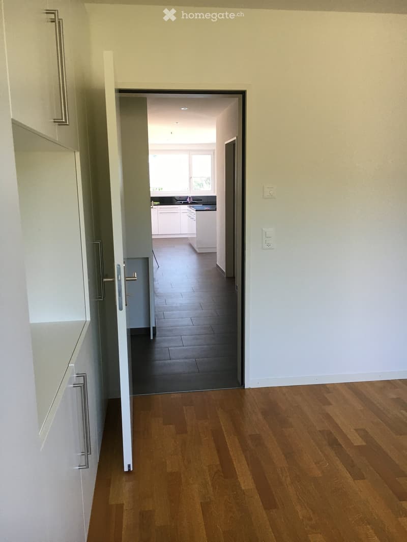 A clean, modern hallway with wooden flooring, white walls, and a door leading to a kitchen area with white cabinets and a dark countertop.