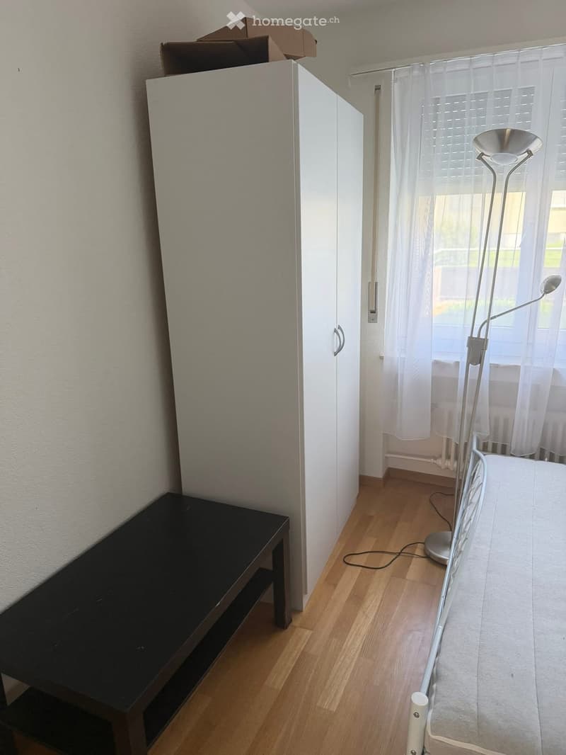 A small, furnished room with a wooden floor, a black table, a white wardrobe, and a floor lamp near a window with sheer curtains.
