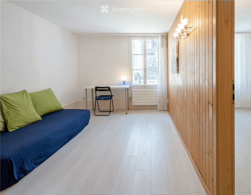 A small, modern bedroom with a blue sofa bed, a small desk with a chair, and a lamp on the desk. The room features light wooden walls and a large window with white curtains.
