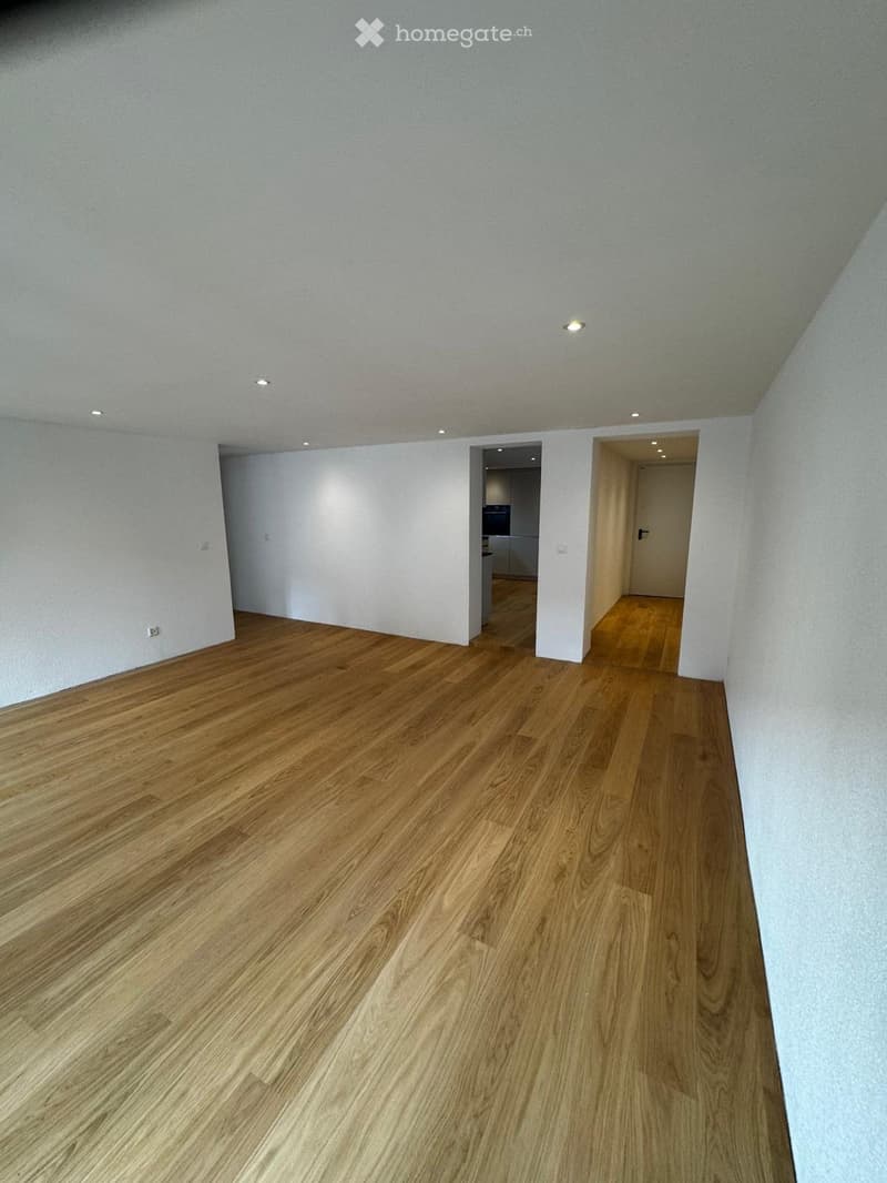 Empty room with light wooden flooring and white walls, featuring recessed ceiling lights and open doorways leading to other rooms.