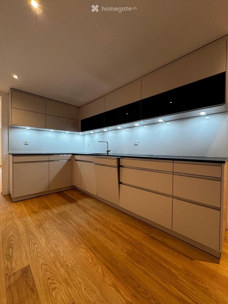 Modern kitchen with light wood cabinets, stainless steel accents, and a sleek black countertop. Features built-in lighting and a minimalist design.