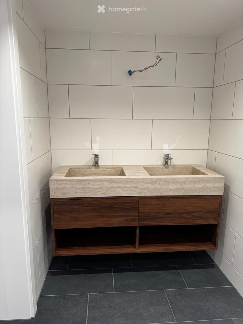 Modern bathroom with a double sink vanity, wooden cabinet, and sleek faucets. The walls are tiled in light gray with a subtle pattern, and the floor is covered in dark gray tiles.