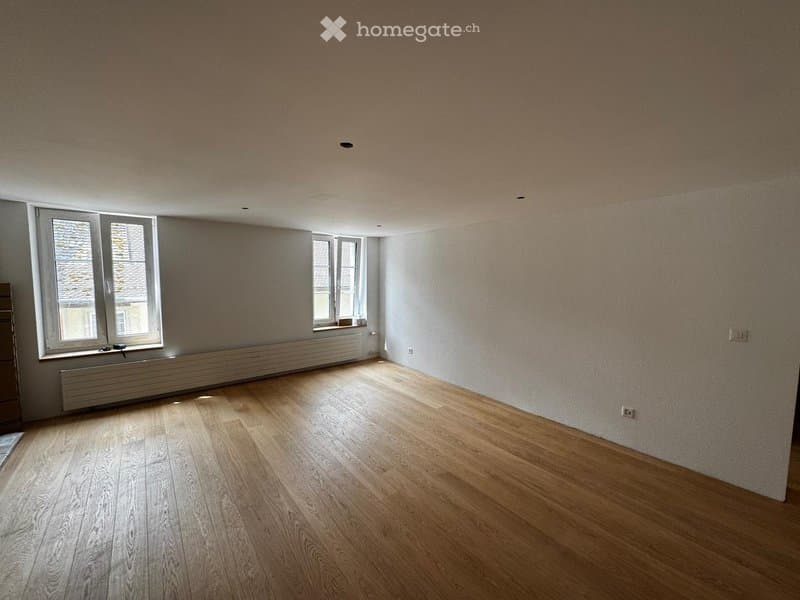 Empty room with wooden flooring, white walls, and two windows with white frames. The room has a radiator under one window and visible electrical outlets on the walls.