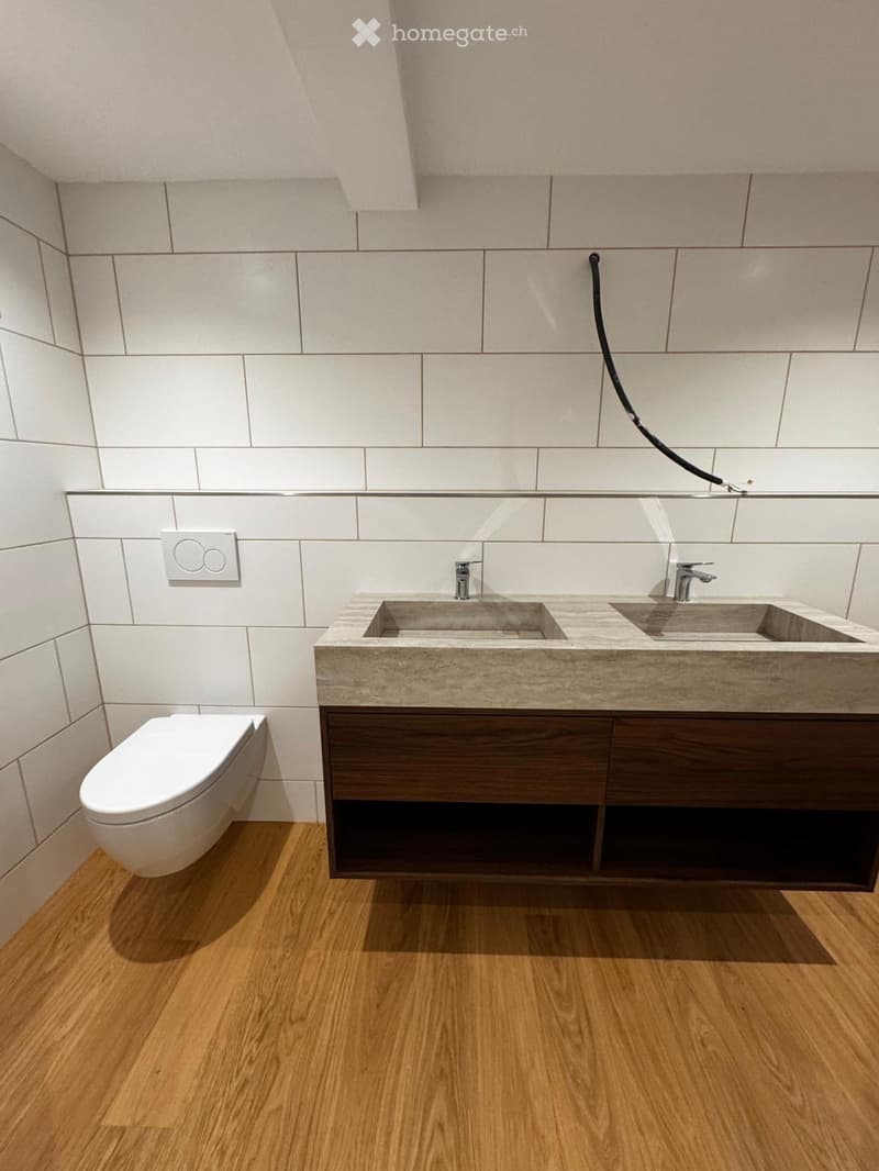 Modern bathroom with a double sink vanity, wooden cabinet, and a toilet. The walls are tiled in a light gray color, and the floor is made of light wood.