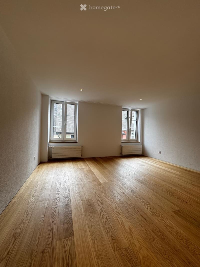 Empty room with wooden flooring, two windows, radiators, and recessed lighting.