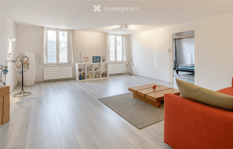 Modern living room with light wooden flooring, white walls, large windows with white curtains, a red sofa, a wooden coffee table, a shelf with decorative items, and a small rug.