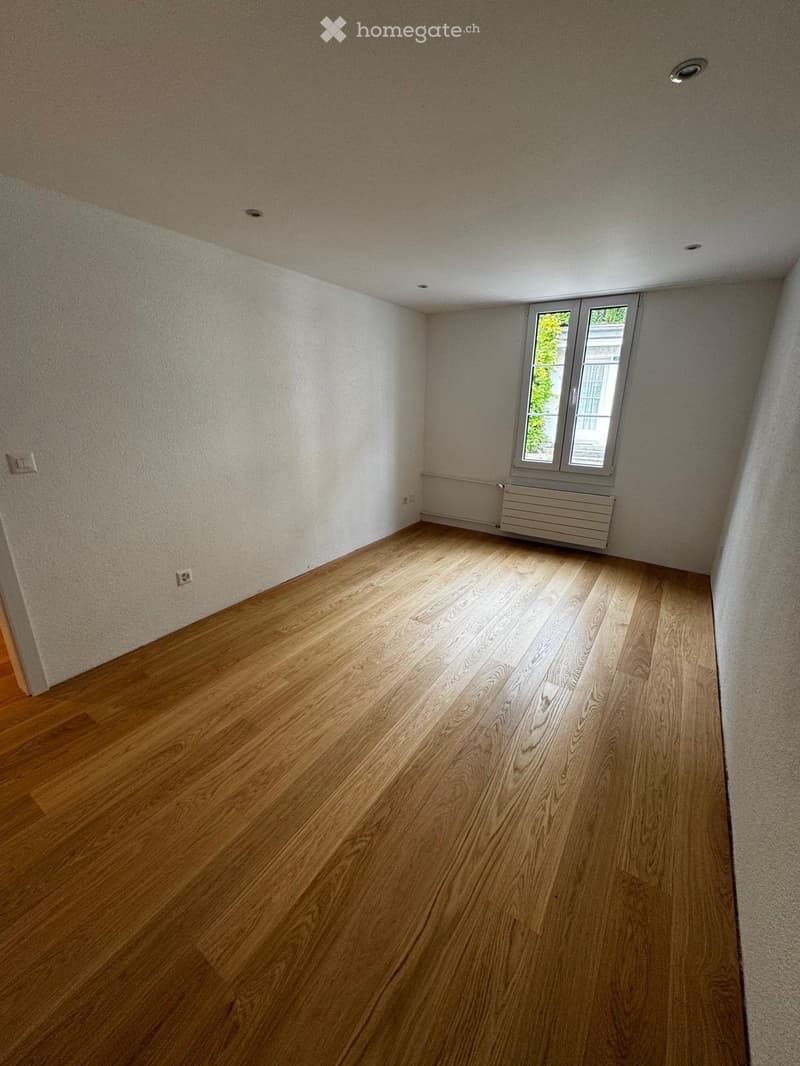 Empty room with wooden flooring, white walls, and a window with a radiator below it.