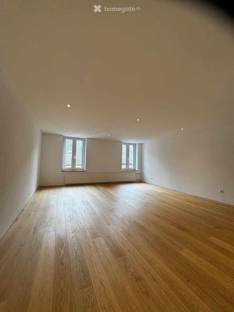 Empty room with wooden flooring, white walls, and two windows with white frames. The room has recessed ceiling lights and a radiator below one of the windows.