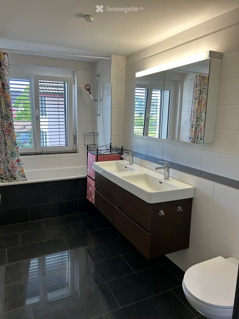 Modern bathroom with dark tiled floor, white sink with dual faucets, large mirror, bathtub with floral shower curtain, and toilet.