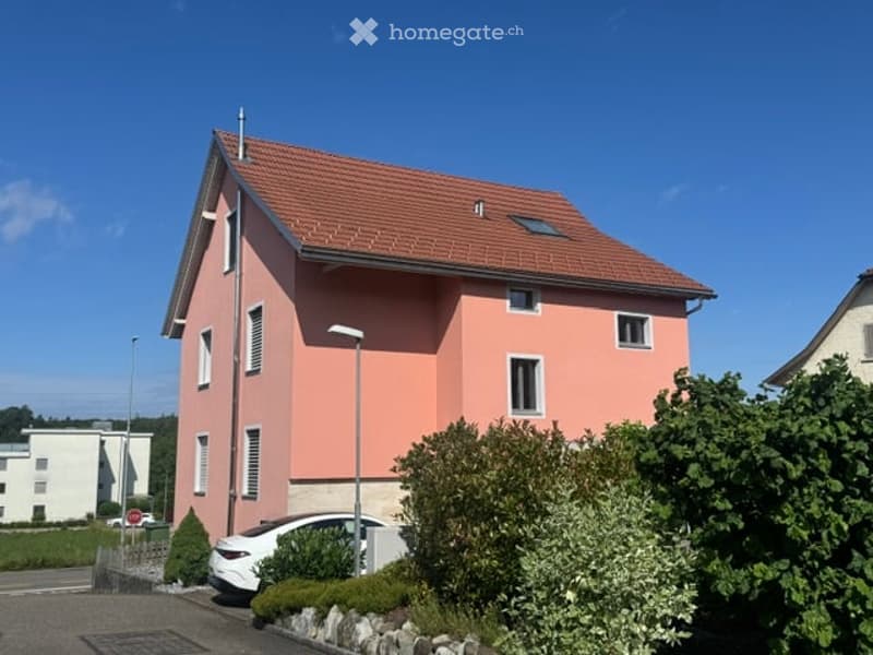 Two-story house with pink exterior, red tile roof, white-framed windows, and a small garden area with a parked car.