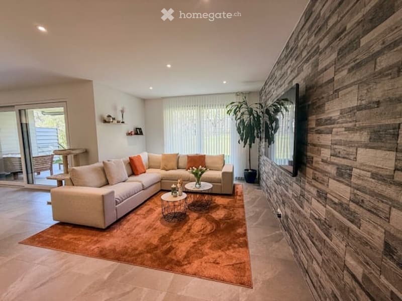 Modern living room with a large sectional sofa, coffee tables, side tables, a television mounted on a textured wooden wall, and a potted plant near the sliding glass door leading to a balcony.