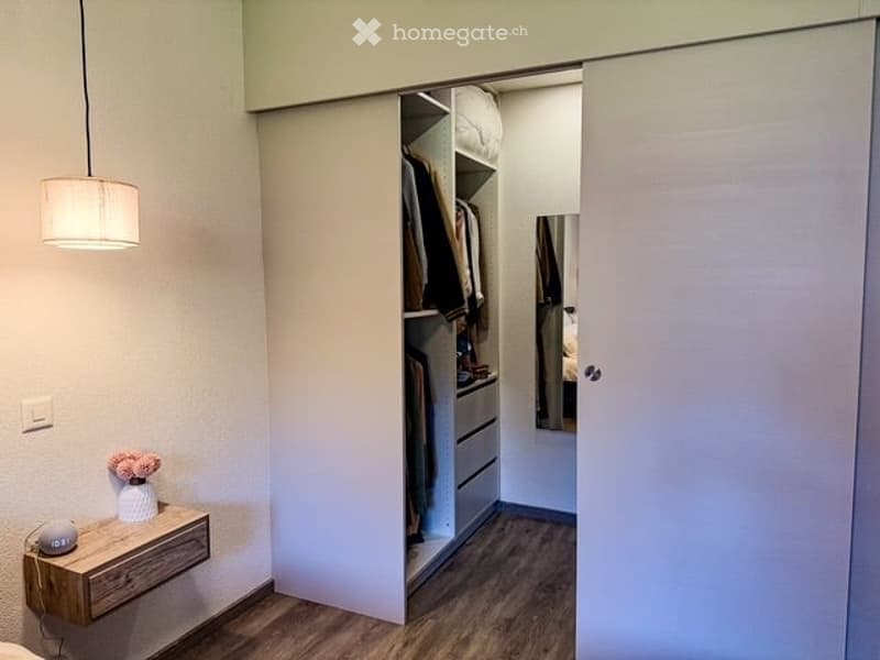 Modern bedroom with a large, open wardrobe, a nightstand with a lamp, and a small decorative vase on the nightstand.
