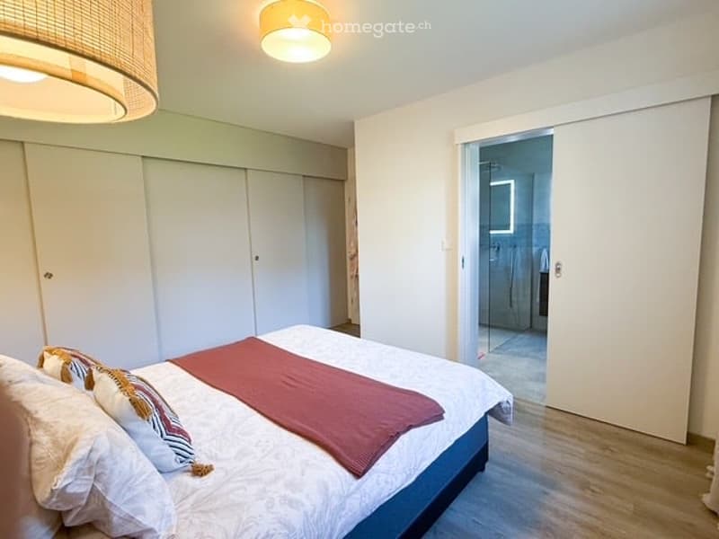 Modern bedroom with a large bed, pillows, a red blanket, and a white wardrobe. The room features light-colored walls and wooden flooring, with a view into an adjacent bathroom.