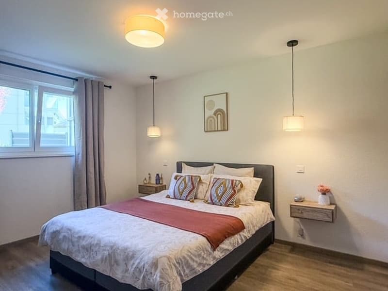 Modern bedroom with a large bed, nightstands, lamps, and a picture frame on the wall. The room features wooden flooring and large windows with curtains.