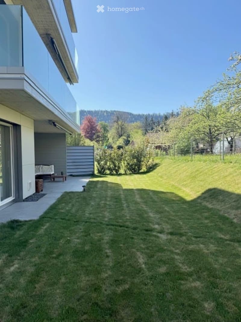 Well-maintained lawn with trimmed grass, surrounded by trees and shrubs, with a modern house and patio visible in the background.