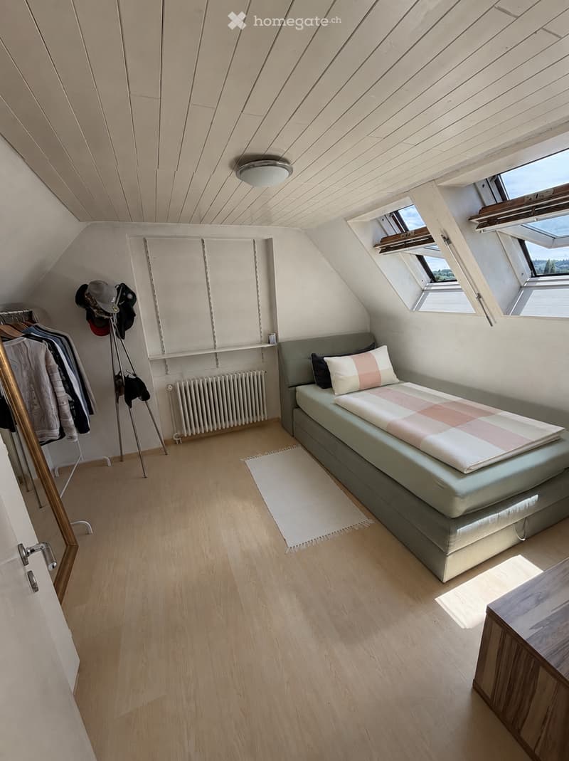 Bright attic bedroom with wooden floors, slanted ceilings, large windows, a single bed with pillows and a blanket, a radiator, a clothing rack with hats, and a small table.