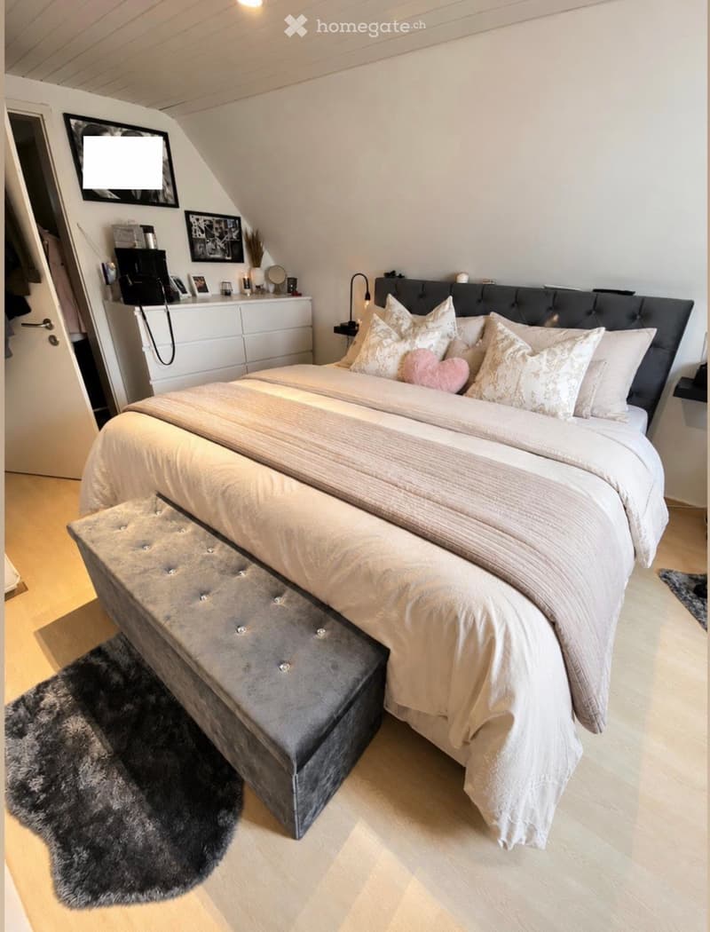 Cozy bedroom with a large bed, bench at the foot, decorative pillows, a mirror, and a small table with a lamp.
