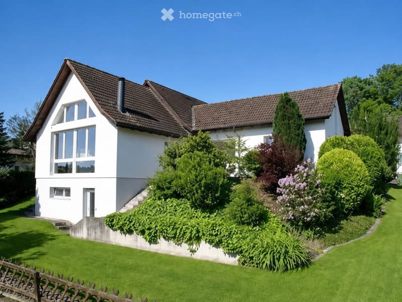 Two-story white house with a brown tiled roof, large windows, and a well-maintained garden with green lawn and various shrubs and trees.