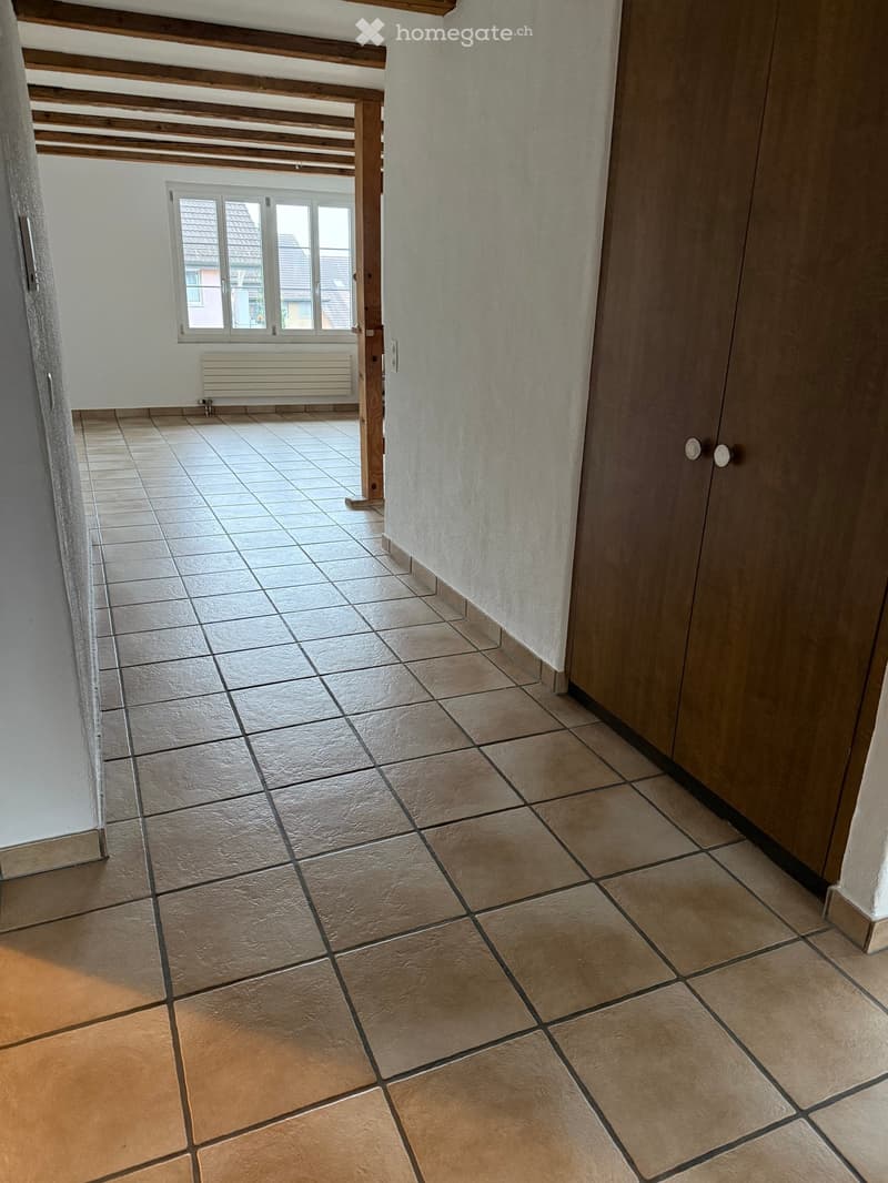 Empty hallway with tiled floor, white walls, wooden beams on the ceiling, and a window at the end.