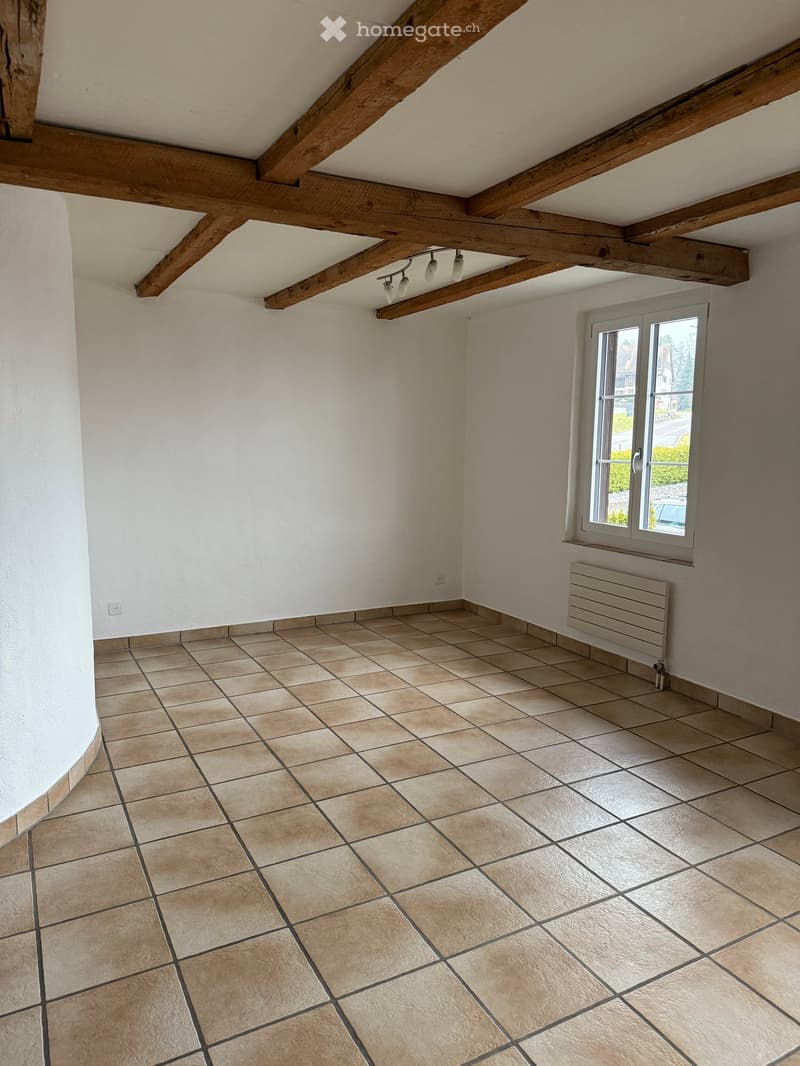 Empty room with exposed wooden beams on the ceiling, tiled floor, white walls, and a window with a radiator below it.